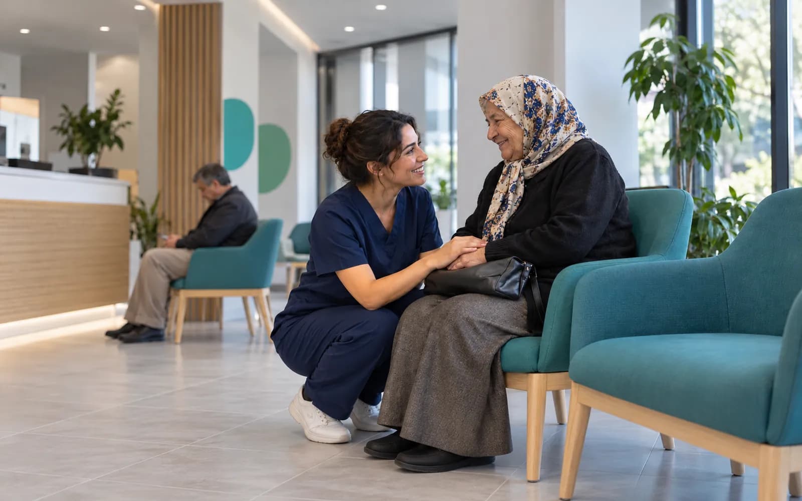 A nurse kneeling to speak with an elderly patient in a waiting area with teal chairs.