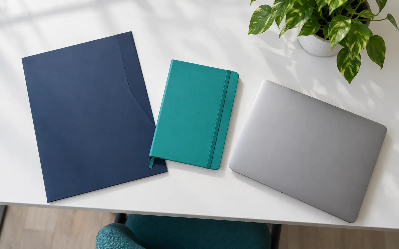Lacivert klasör, turkuaz defter, kapalı dizüstü ve bitki bulunan, yukarıdan çekilmiş sade bir basın kiti masası